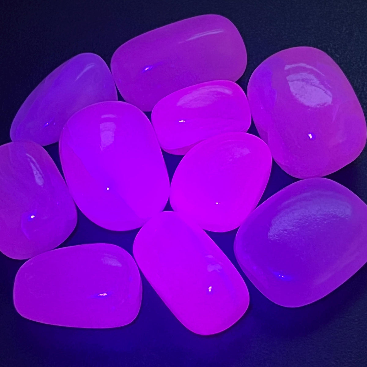 Pink mangano calcite tumbled stones fluorescing vivid pink and purple under UV light on a dark surface.