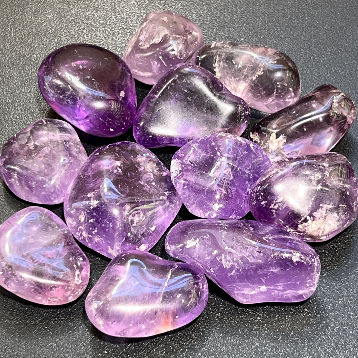 Polished amethyst tumbled stones in lavender and deep purple with rounded shapes on a dark textured surface.