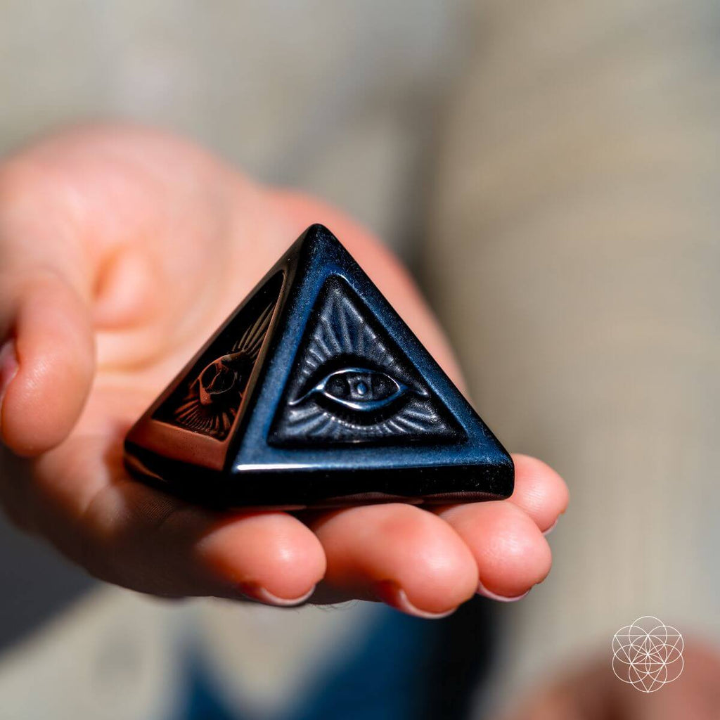 Polished black obsidian pyramid with carved all-seeing eye designs held in an open hand.
