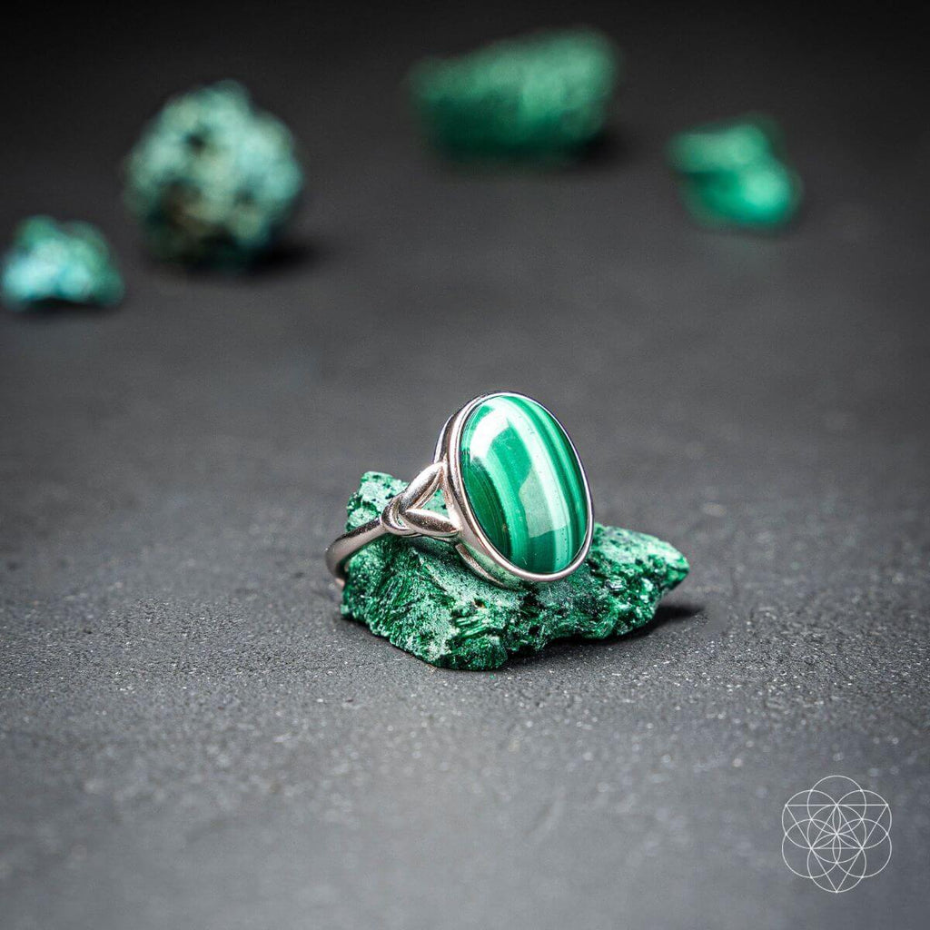 Polished oval malachite ring resting on a rough green stone against a dark background.

