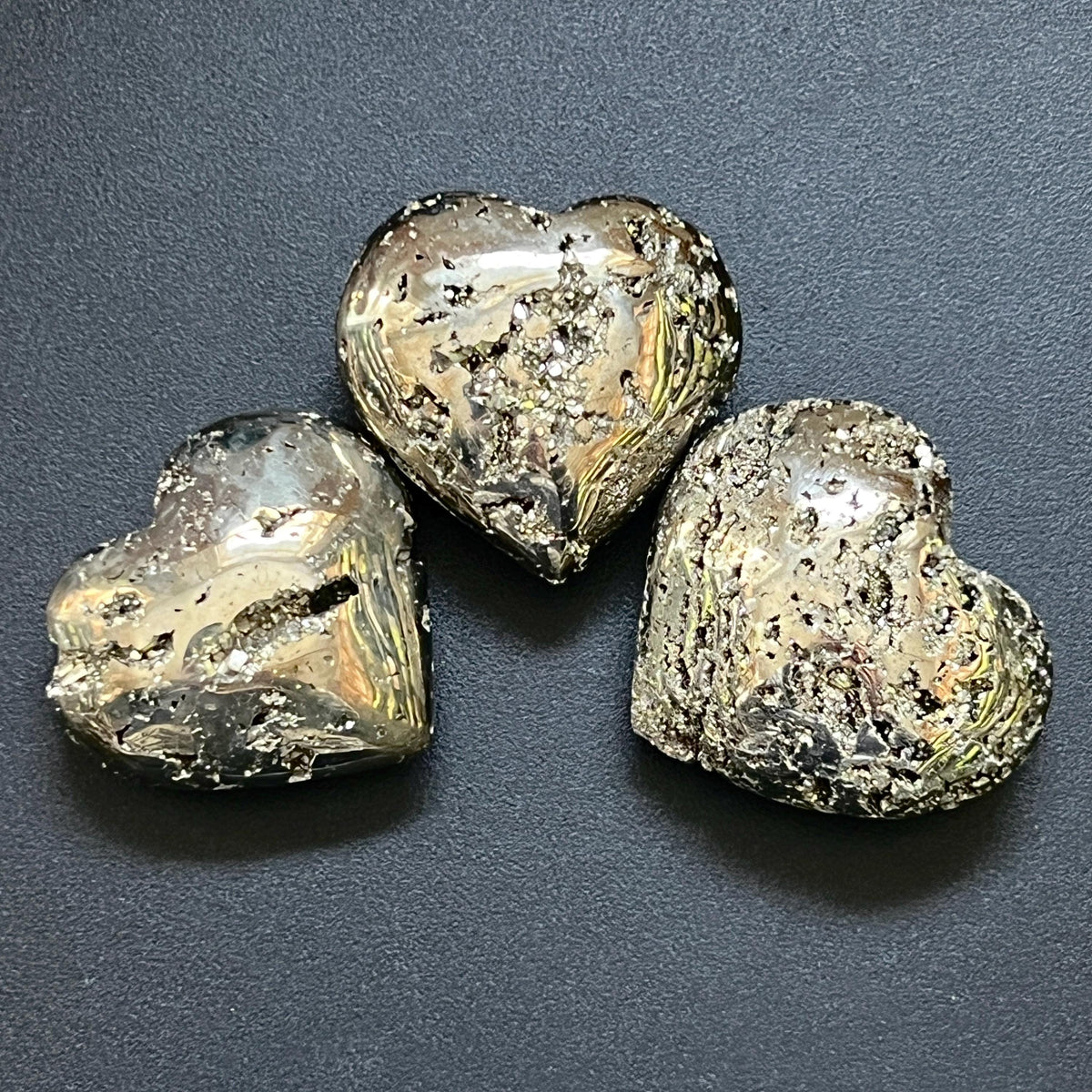 Polished pyrite heart carvings with reflective metallic faces and irregular crystal pockets arranged on a dark surface.