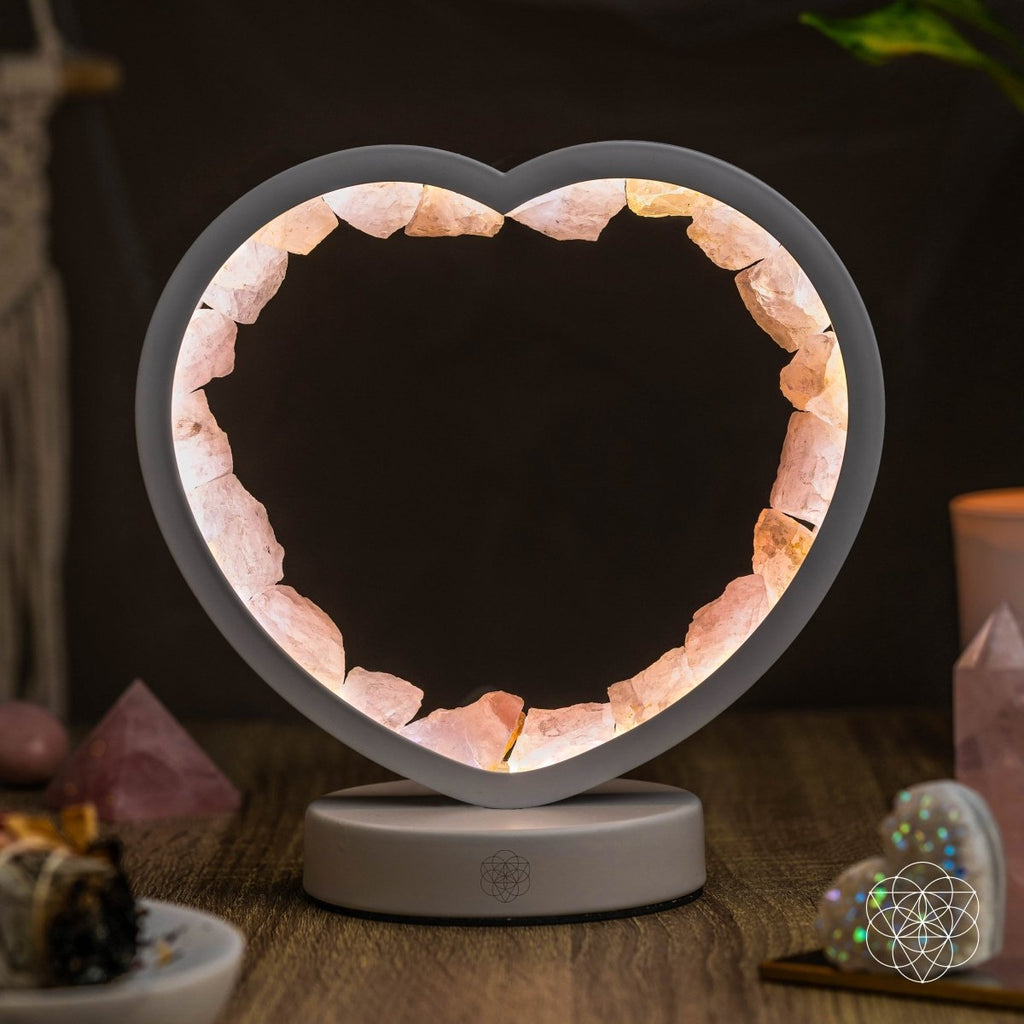 Rose quartz heart lamp lit against a dark background with decorative crystals and a candle nearby.

