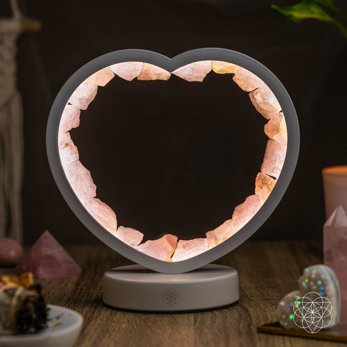 Rose quartz heart lamp lit against a dark background with decorative crystals and a candle nearby.
