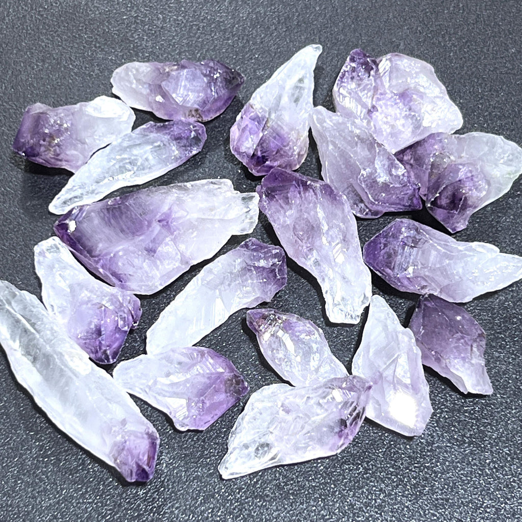 Rough amethyst crystal points with pale purple tips and translucent white sections on a dark textured surface.
