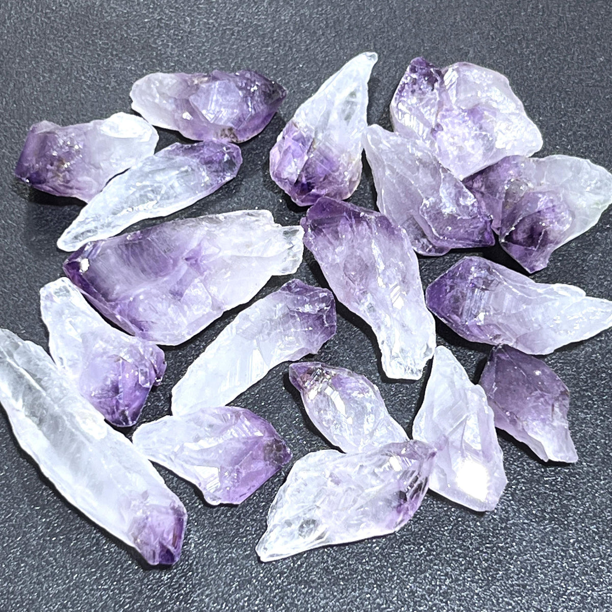 Rough amethyst crystal points with pale purple tips and translucent white sections on a dark textured surface.
