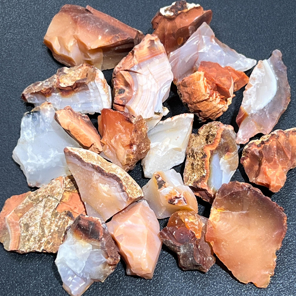 Rough carnelian agate stones with red-orange and milky white areas arranged on a dark textured surface.