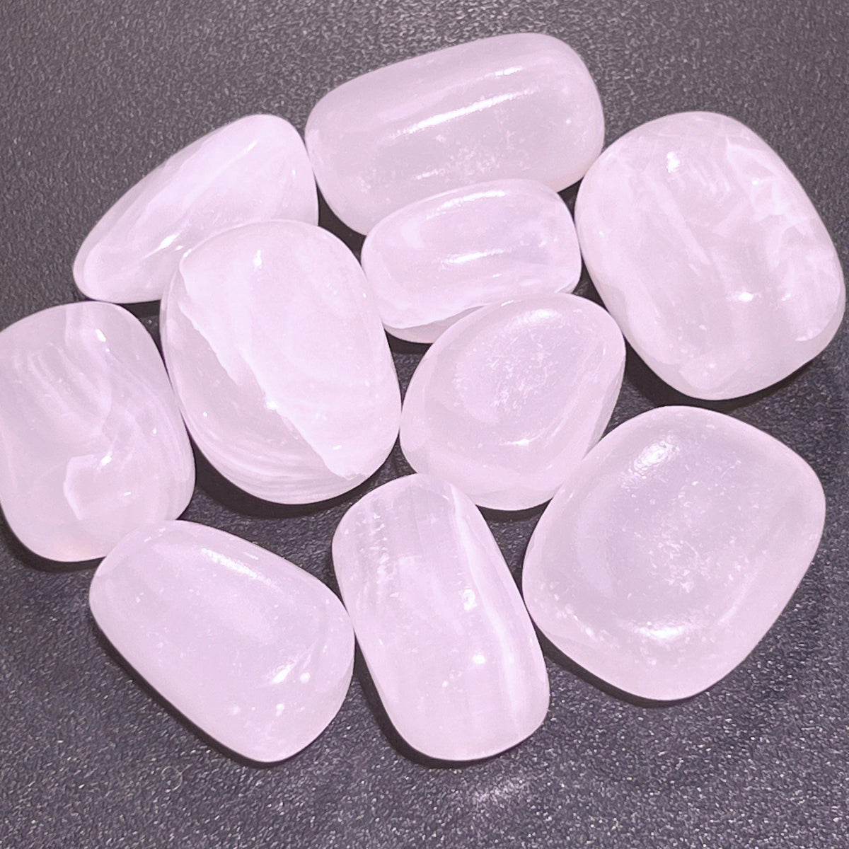 Rounded pink mangano calcite tumbled stones with pale banding arranged on a dark textured surface.