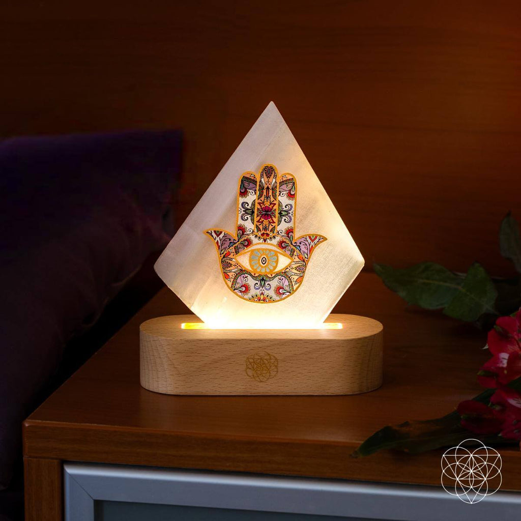 Selenite Hamsa hand lamp casting a warm glow on a wooden bedside table beside red flowers
