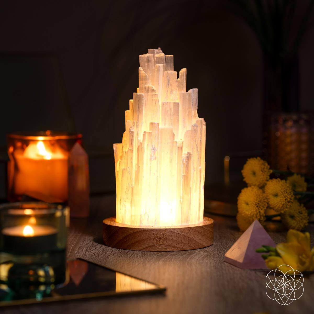Selenite tower lamp casting a warm glow on a tabletop beside candles, yellow flowers, and a pale pyramid.
