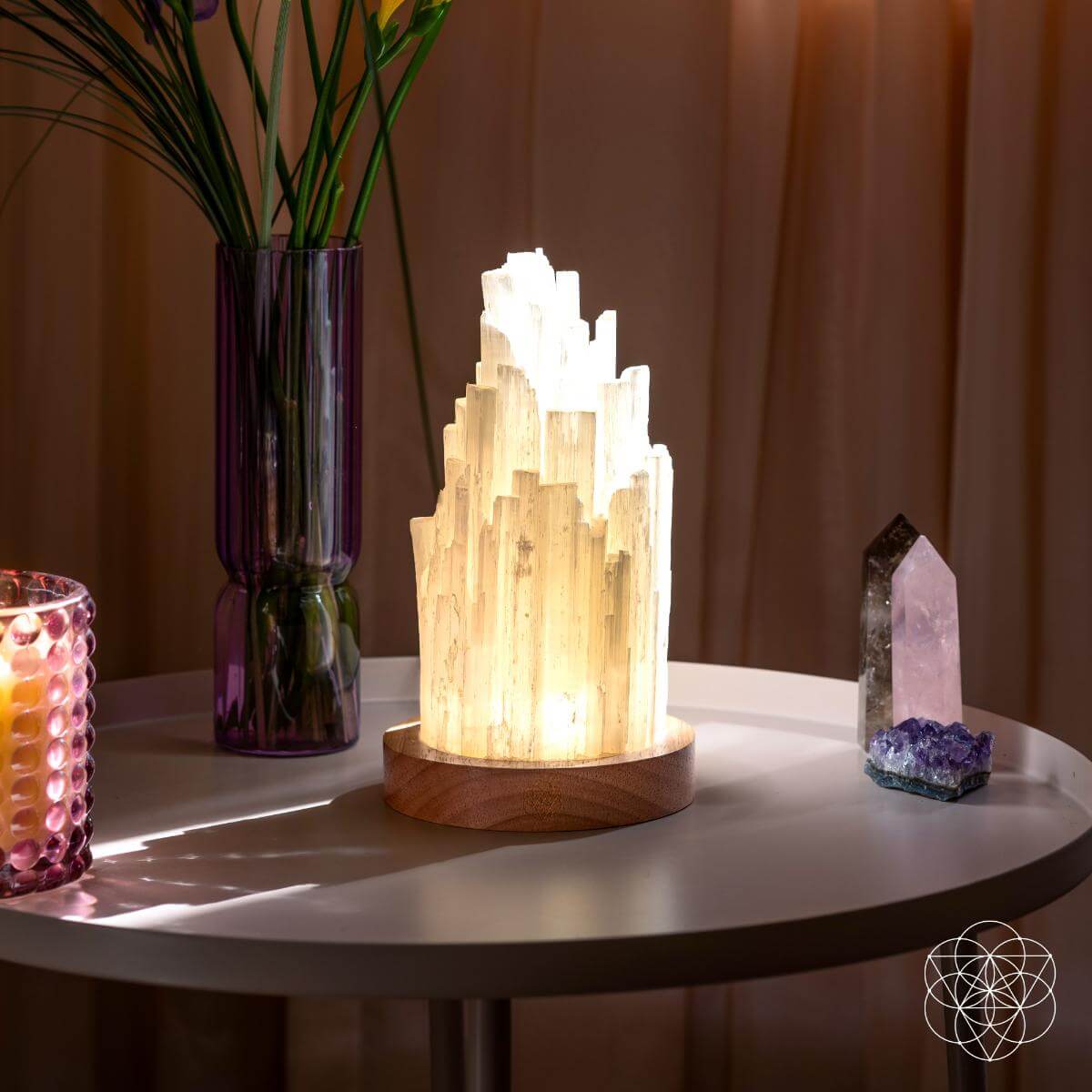Selenite tower lamp on a round table beside a tall purple vase, pink candle, and crystal points.
