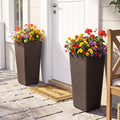 Two tall dark brown wicker-style planter boxes filled with colorful flowers, placed on either side of a white front door on a stone walkway. A tan doormat with the word “WELCOME” is visible in front.