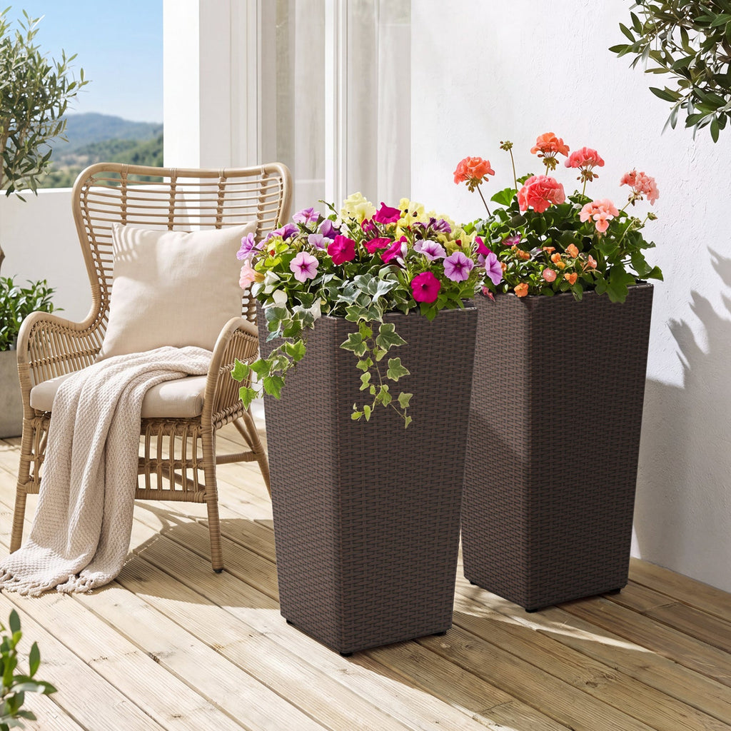 Two tall rectangular dark brown wicker-style planter boxes filled with colorful flowers, placed on a wooden deck beside a wicker chair with a cream throw blanket.