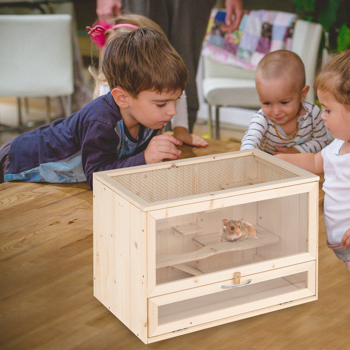 A light natural wood hamster cage with glass panels and a small hamster inside, shown on a wooden table with children looking at it in a home setting.
