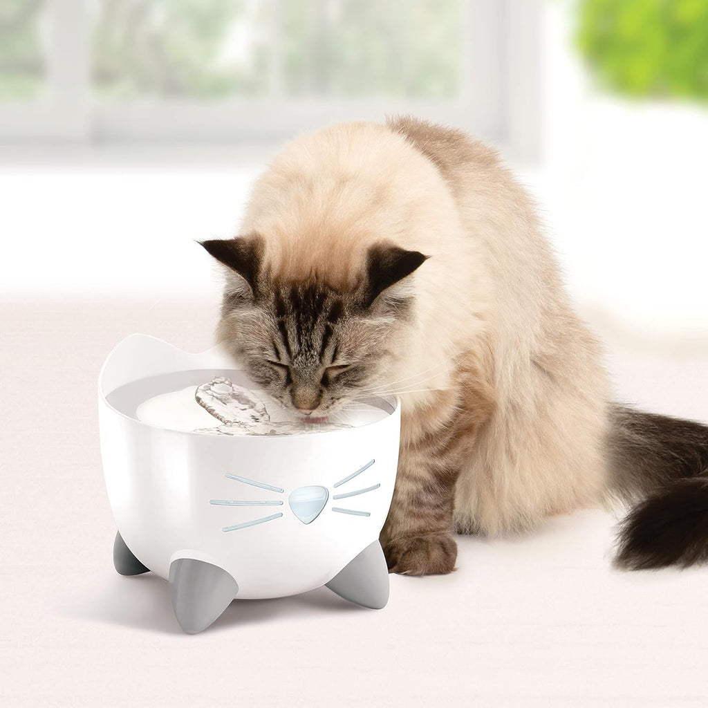 Cat drinking water from a white cat-shaped water fountain on a light surface.