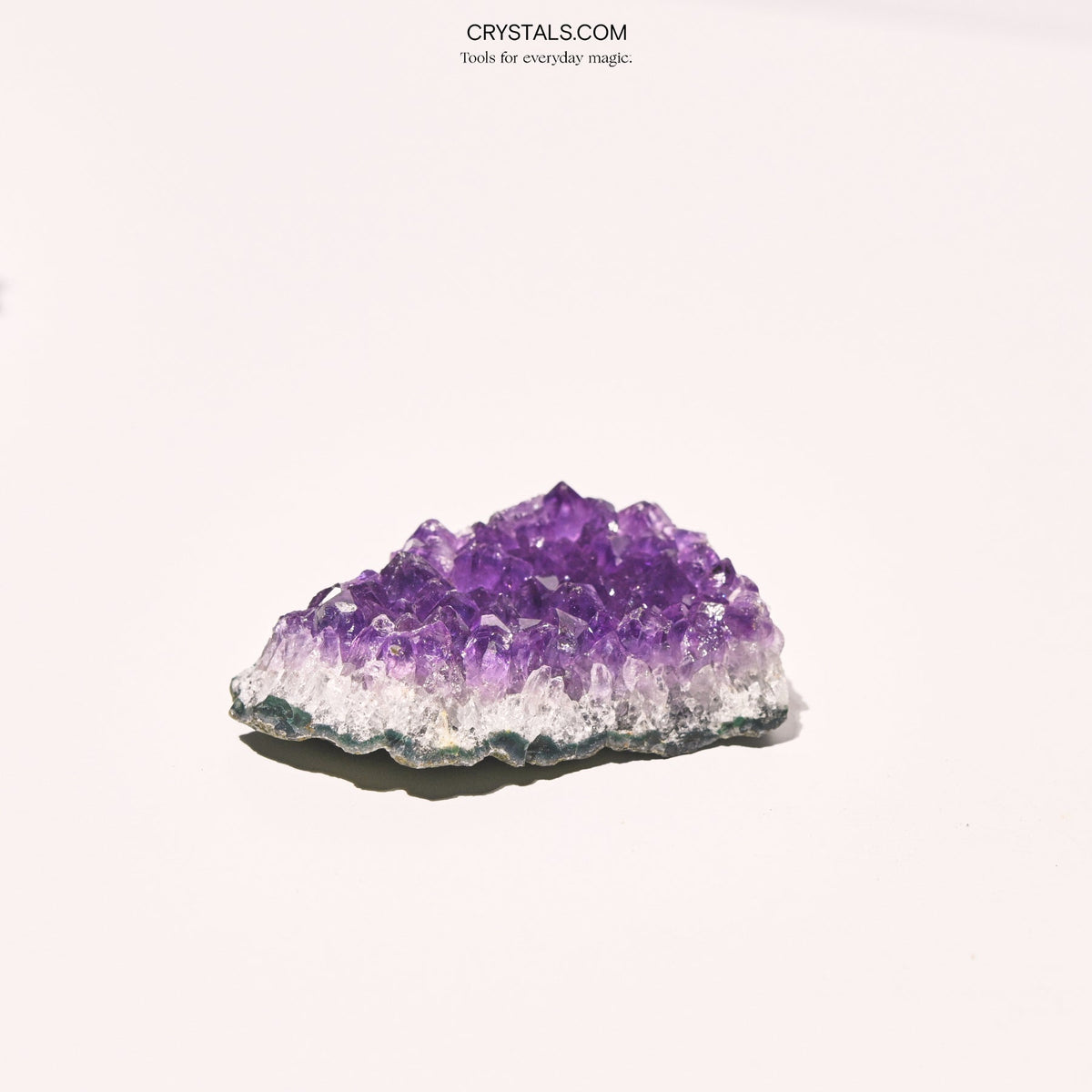 Side view of a low amethyst cluster showing purple crystal points above a pale band and dark base.
