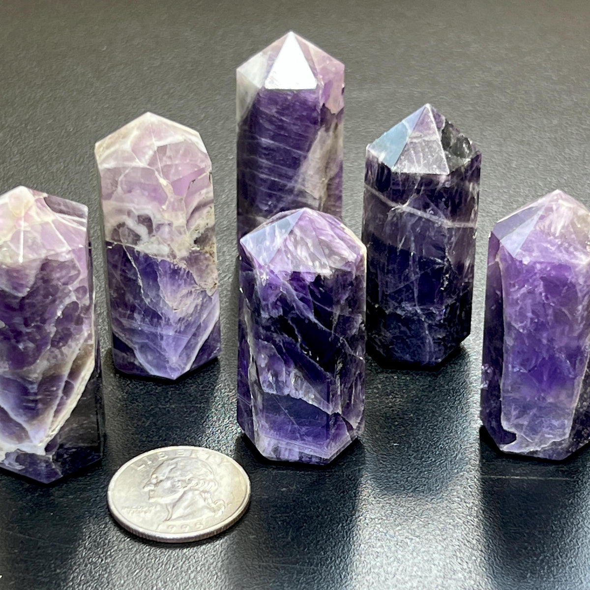 Six polished banded amethyst crystal towers with pointed tops on a dark surface beside a quarter for scale
