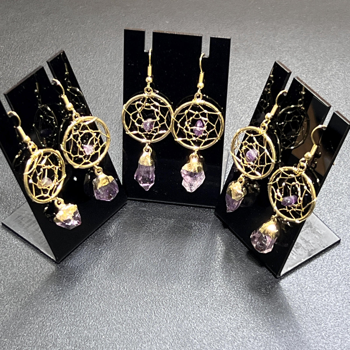 Three pairs of gold-plated amethyst dreamcatcher dangle earrings displayed on black earring stands.
