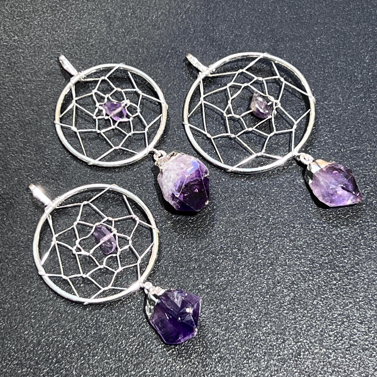 Three silver-plated amethyst dreamcatcher pendants on a dark textured surface showing varied hanging crystal shapes