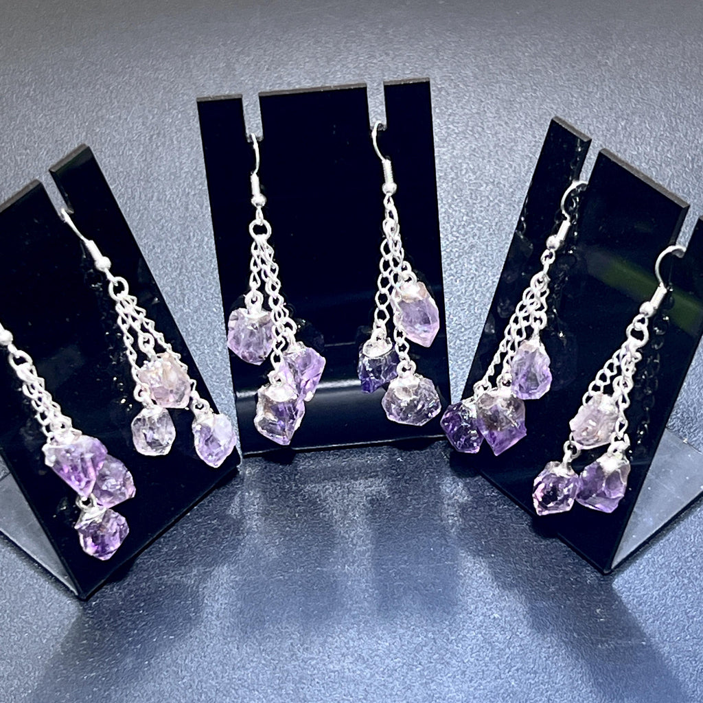 Three pairs of purple amethyst drop earrings with silver-colored hooks and staggered chains on black stands.