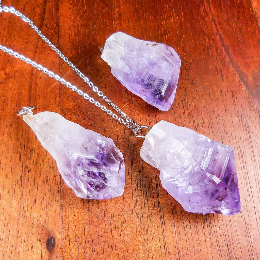 Three raw purple amethyst pendants on reddish wood, with one pendant styled on a silver-colored chain.
