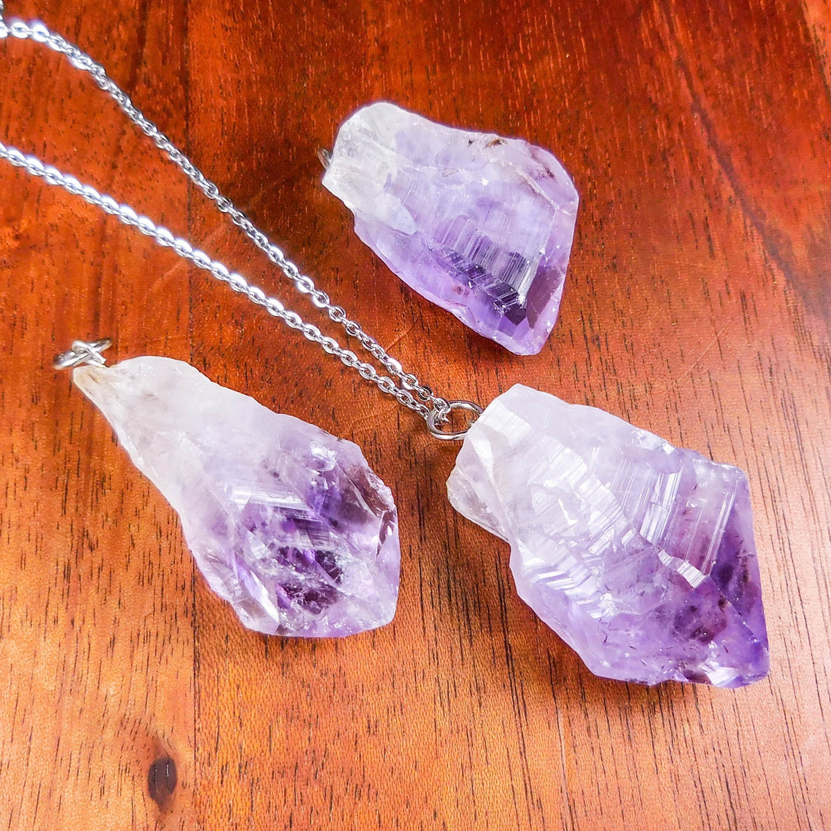 Three raw purple amethyst pendants on reddish wood, with one pendant styled on a silver-colored chain.
