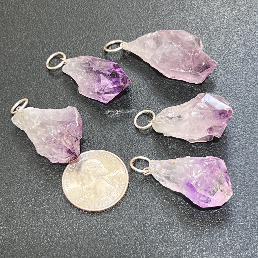 Five raw amethyst point pendants with silver-tone rings beside a quarter for size comparison.
