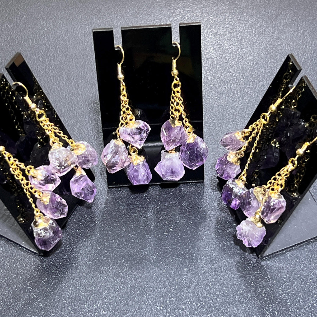 Three pairs of amethyst triple crystal dangle earrings with gold-tone chains and hooks on black display stands.