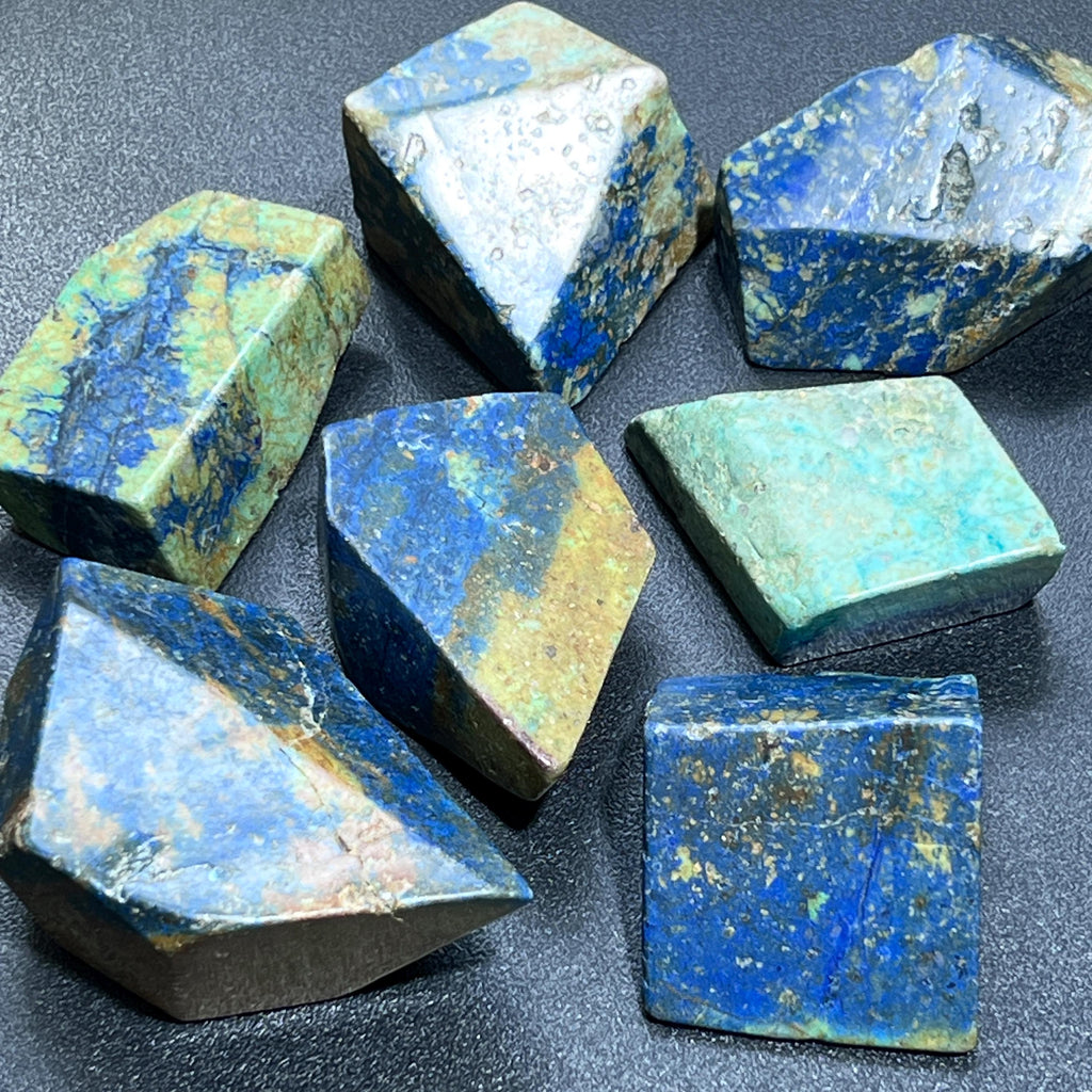 Assorted angular azurite and chrysocolla polished stones showing blue and turquoise-green patterns on a dark surface.