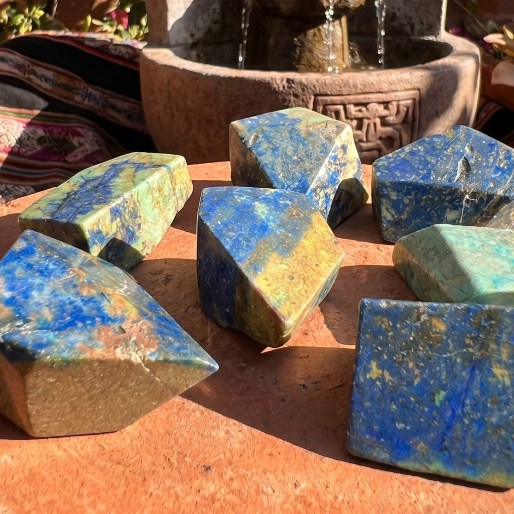 Blue and green azurite and chrysocolla freeform stones on a sunlit terracotta-colored surface near a fountain.