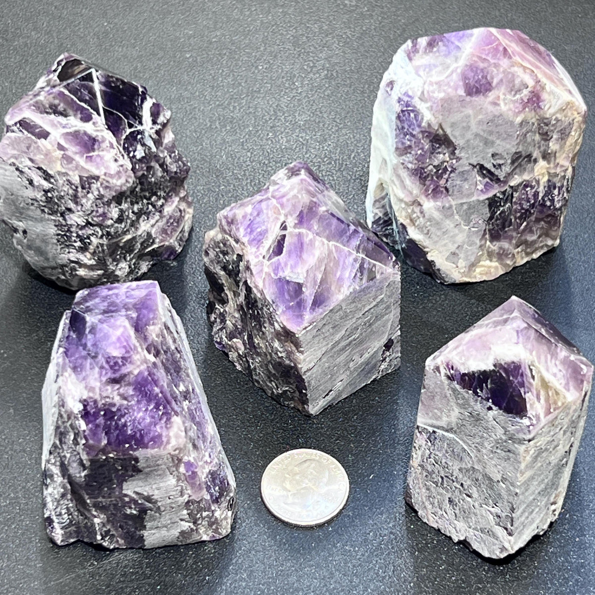 Five purple and white banded amethyst point towers beside a coin for size comparison on a dark surface.
