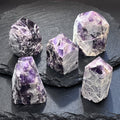 Five banded amethyst towers with polished purple points and rough pale sides arranged on layered dark slate.
