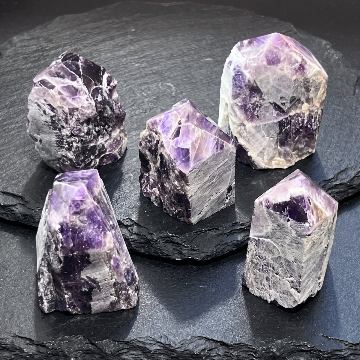 Five banded amethyst towers with polished purple points and rough pale sides arranged on layered dark slate.
