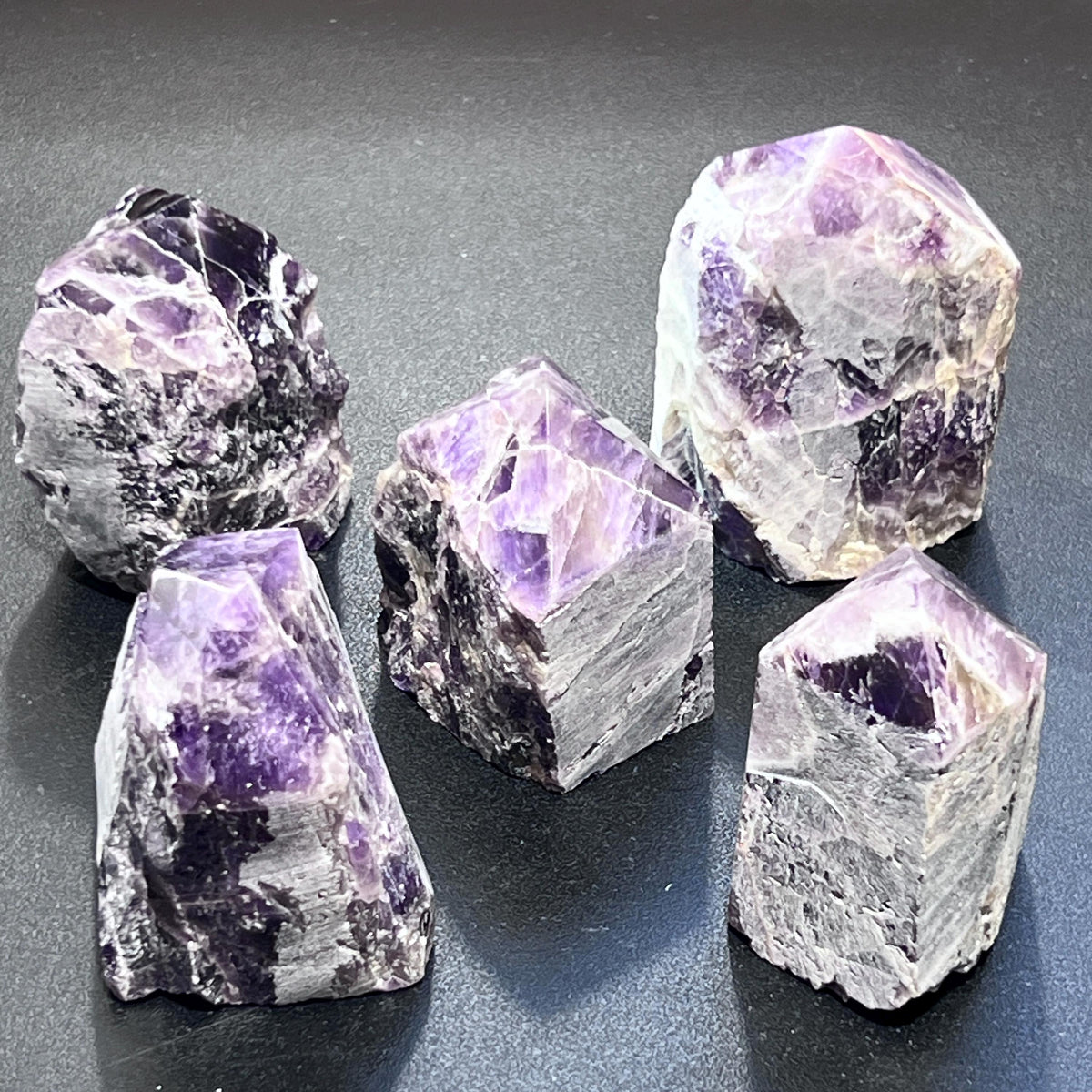 Five standing banded amethyst towers showing polished pointed tops and textured unpolished sides on a dark background.
