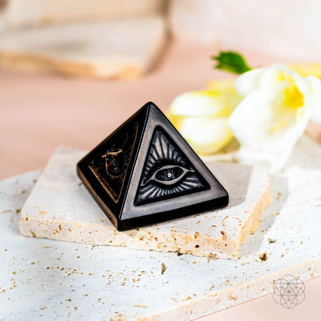 Glossy black obsidian pyramid with an eye and radiating lines carved into its faces on cream stone slabs.
