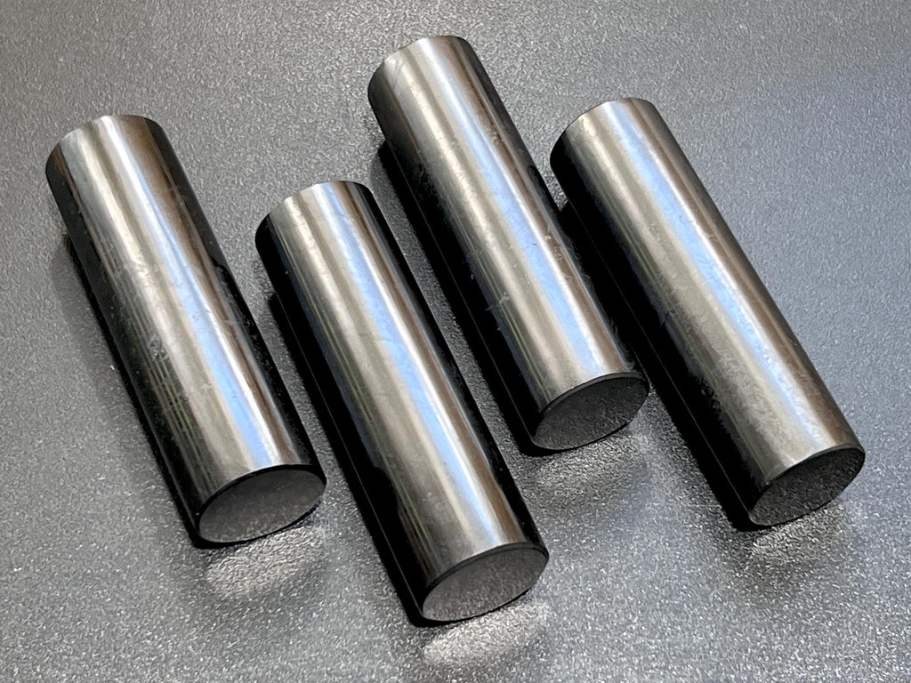 Four polished black shungite cylinder rods with reflective sides and flat circular ends on a dark surface.
