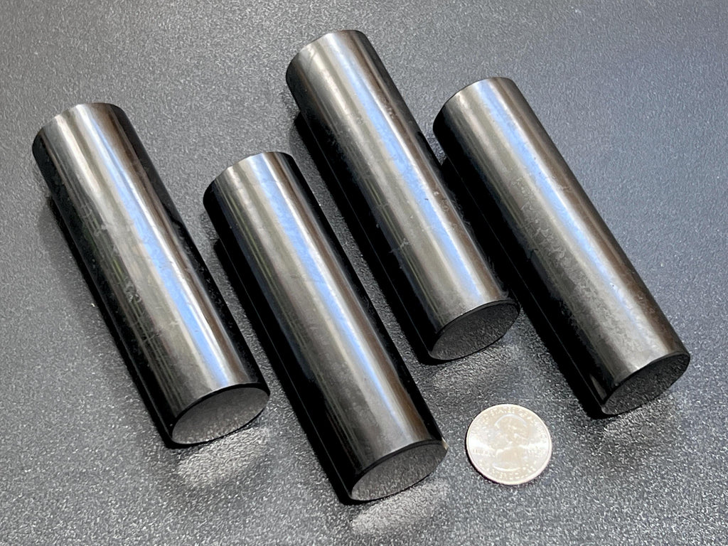 Four polished black shungite cylinder rods beside a quarter for size comparison on a dark textured background.
