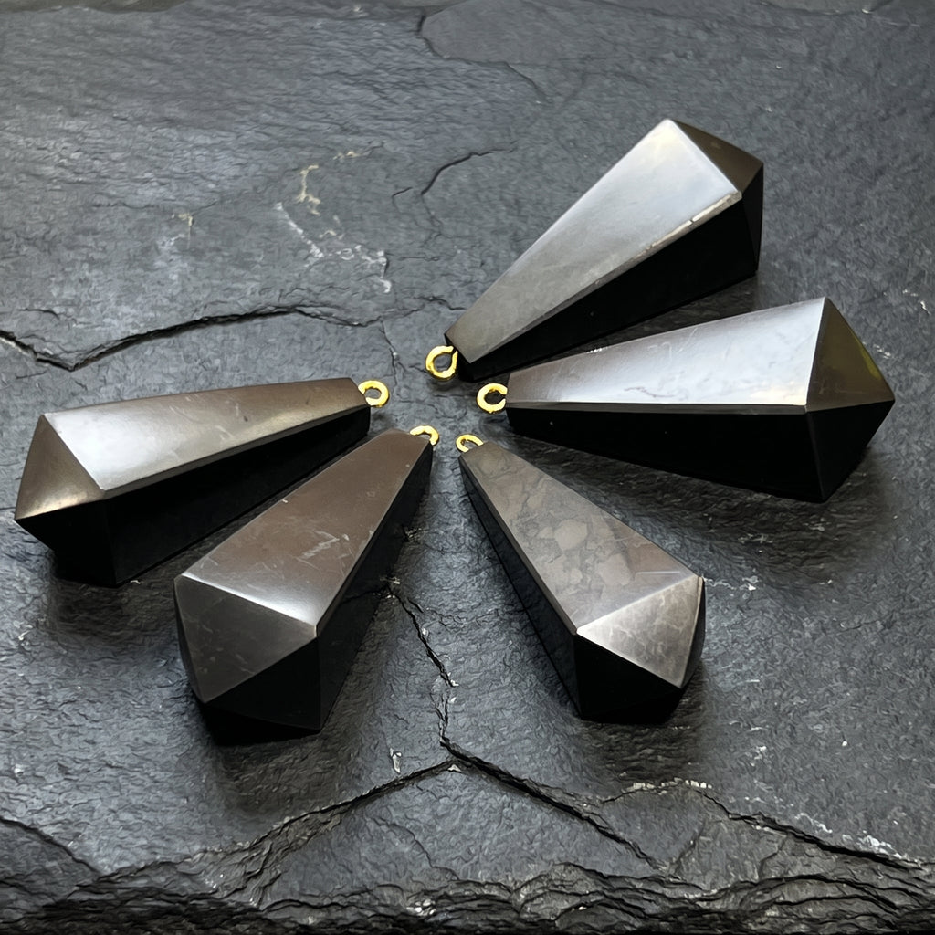 Angled view of five tapered black shungite pendants showing flat facets and small hanging loops on slate.
