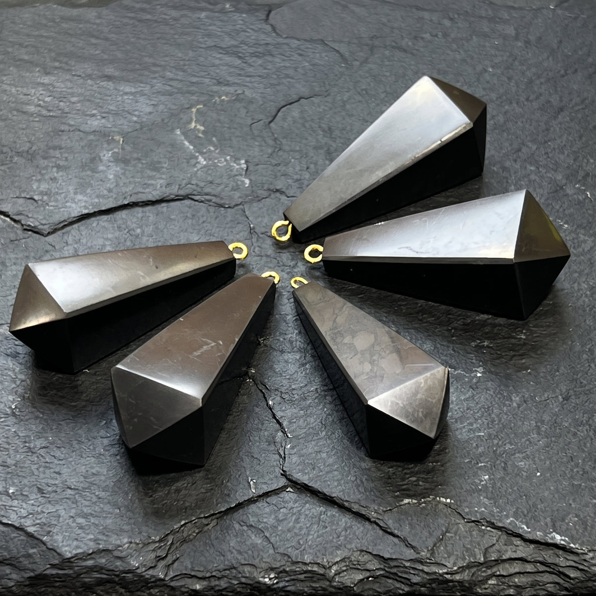 Angled view of five tapered black shungite pendants showing flat facets and small hanging loops on slate.
