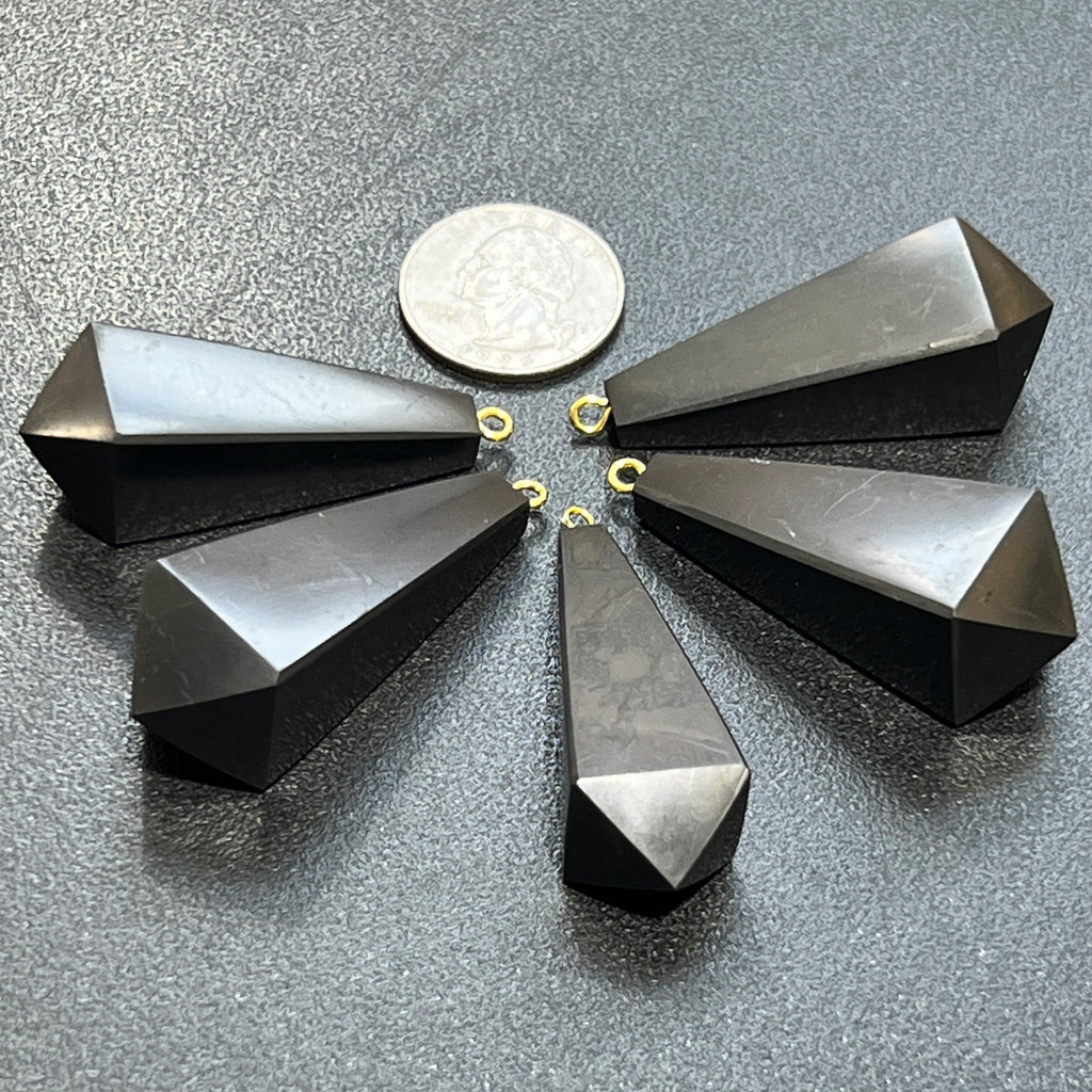 Five faceted black shungite pendulum point pendants with gold-colored hanging loops arranged on textured slate.
