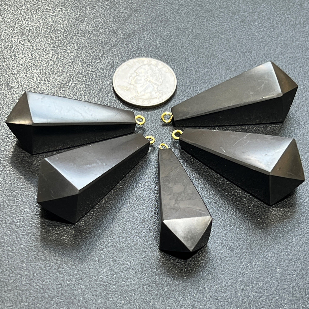 Five faceted black shungite pendulum point pendants with gold-colored hanging loops arranged on textured slate.
