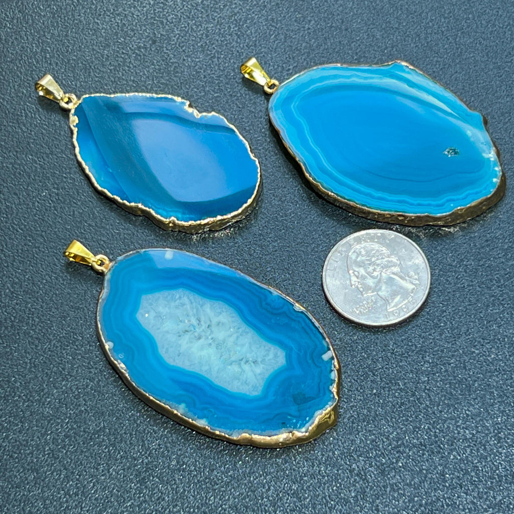 Three blue agate pendants with gold edges on a dark surface, one next to a coin for scale.
