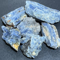 Six rough blue kyanite crystal pieces with layered blade-like formations and pale gray and cream areas on a dark surface
