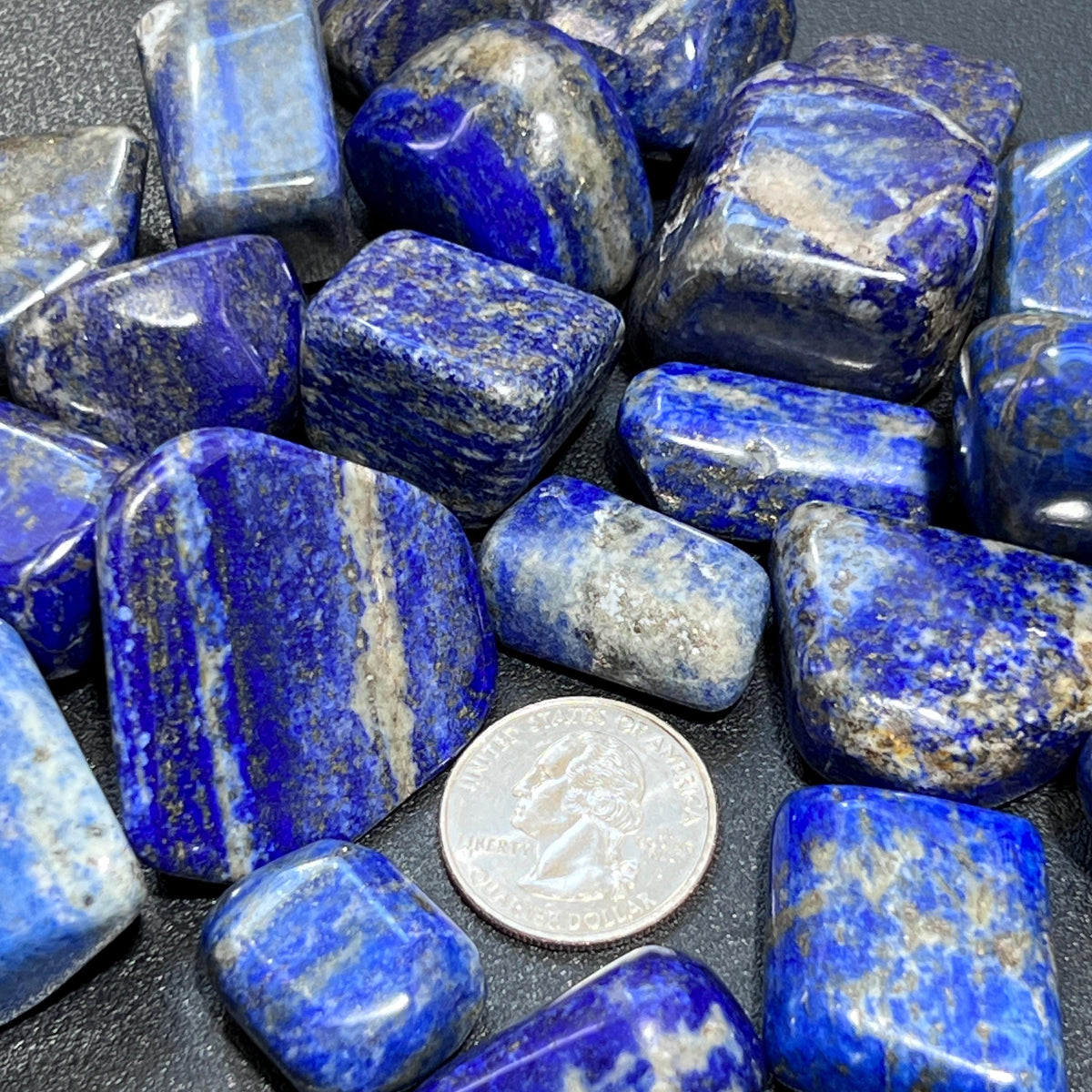 Close-up of blue lapis lazuli tumbled stones with pale markings beside a quarter for size comparison.
