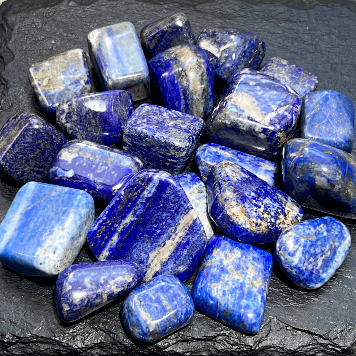 Polished blue lapis lazuli tumbled stones with white veins and gold-colored flecks arranged on dark slate.
