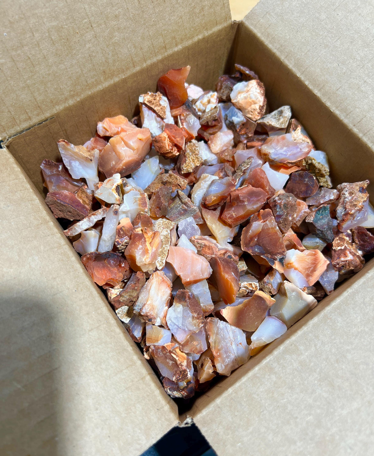 Angled view into a cardboard box filled with rough red, orange, and white carnelian agate pieces.