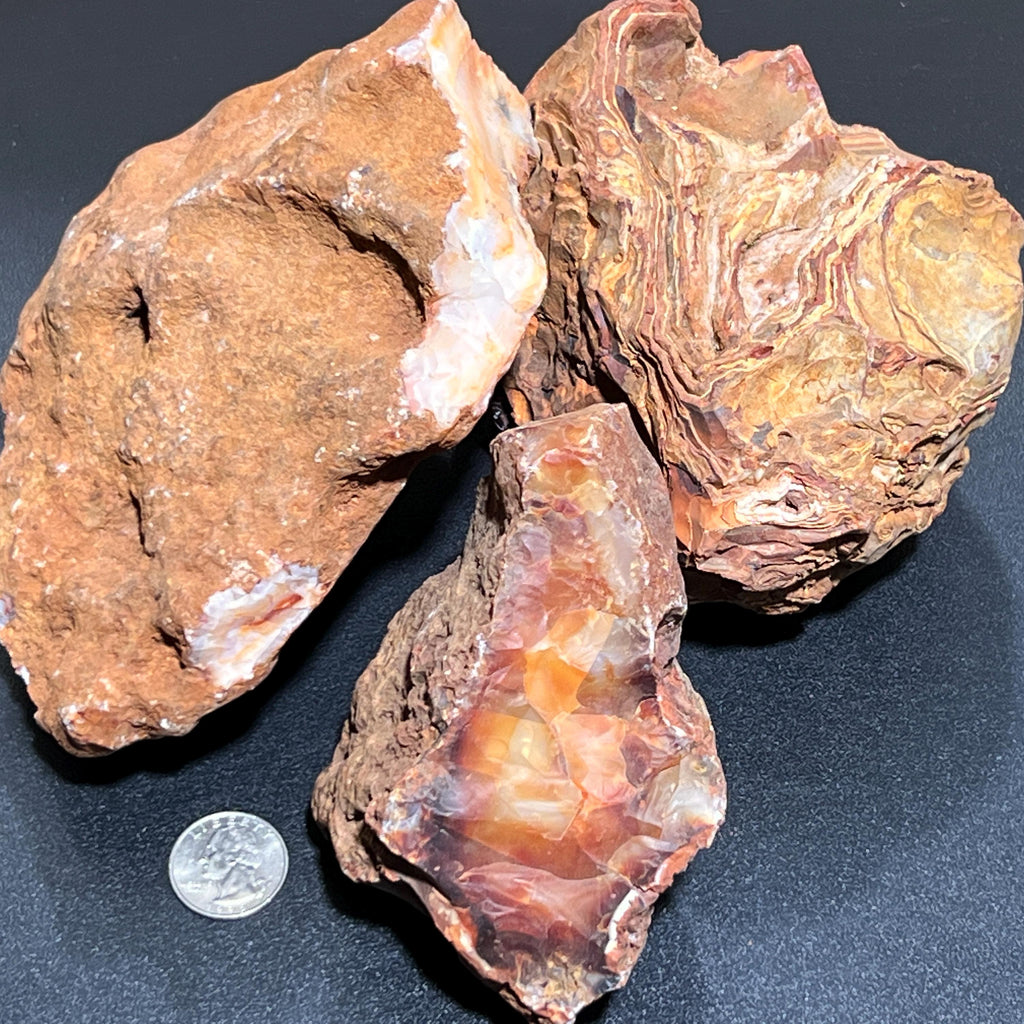 Three rough carnelian agate stones on a dark surface beside a quarter for scale