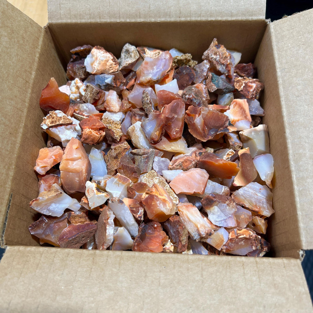 Overhead view of a bulk assortment of rough carnelian agate stones in an open cardboard box.