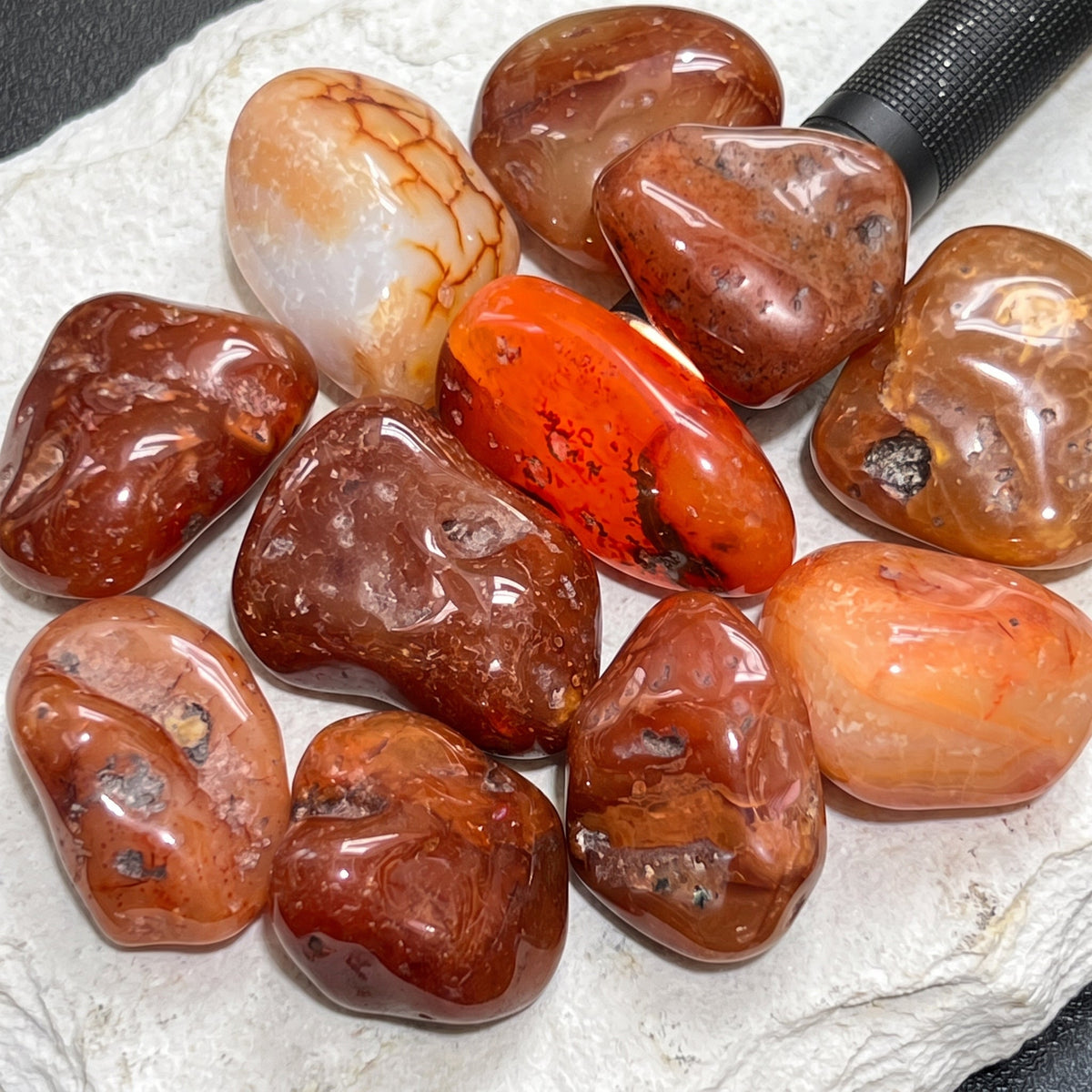 Glossy carnelian red agate stones arranged on a white slab, with a flashlight illuminating an orange stone