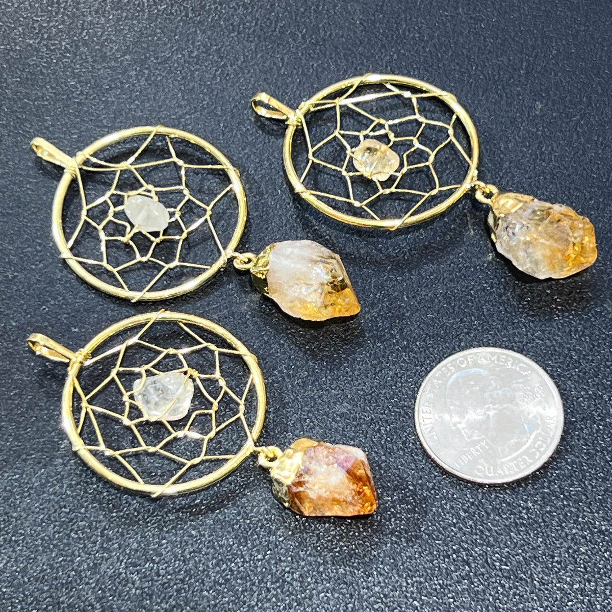 Three gold-plated citrine dreamcatcher pendants beside a quarter for size comparison on a dark surface.
