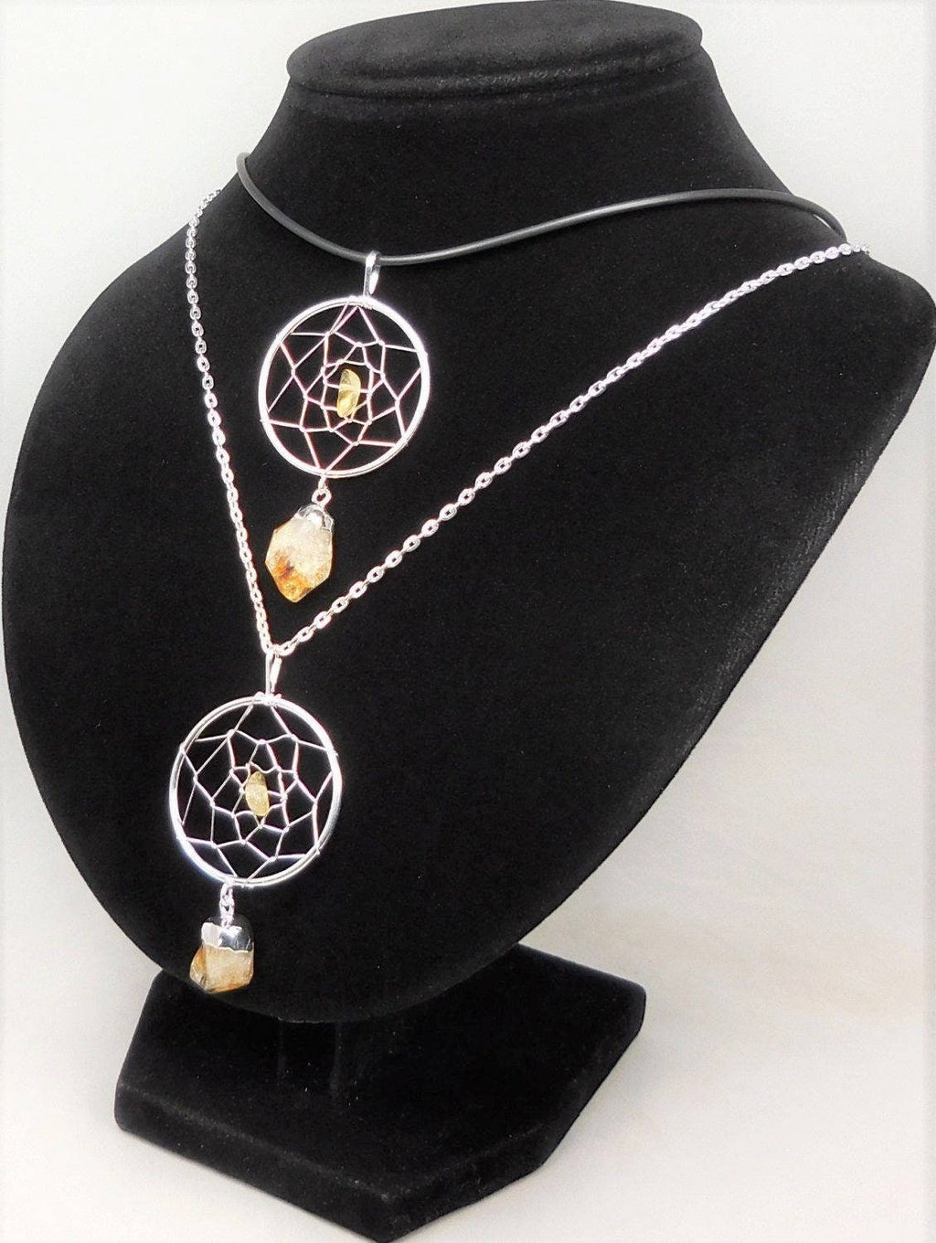 Two silver-plated citrine dreamcatcher pendants styled on a black necklace bust, one on a cord and one on a chain.

